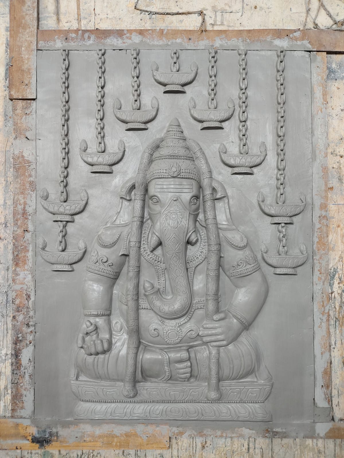 Grey cement relief of a seated Ganesha with hanging oil lamps