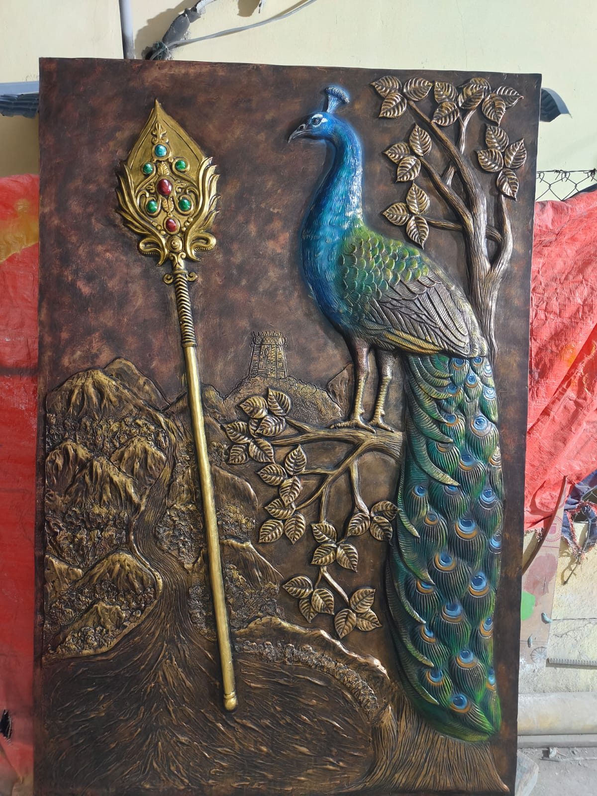 Bronze-finish relief of a peacock beside a sacred vel, with hills and a temple tower